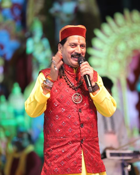 Prem Bhatnagar ji singing on stage in a red jacket and pahadi cap
