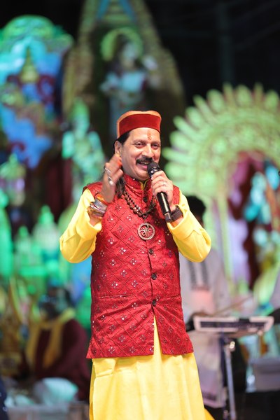 Prem Bhatnagar ji singing on stage in a red jacket and pahadi cap