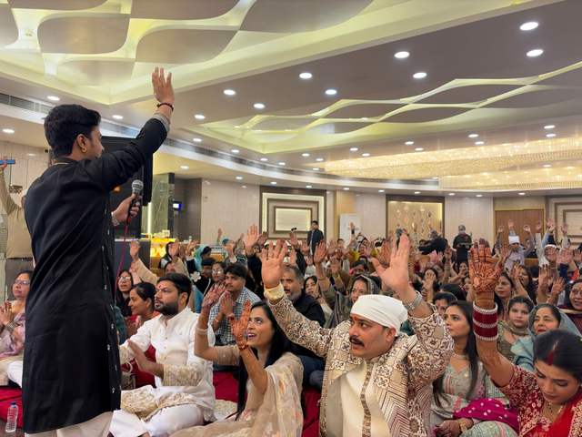 A singer of the party leading a packed hall of devotees, everyone raising their hands to Mata's bhajan