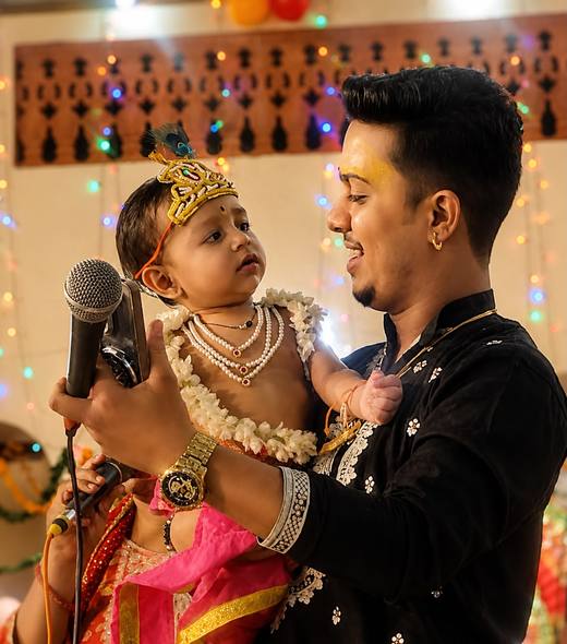 A singer of the party smiling at a baby dressed as Bal Gopal, with a peacock-feather crown and jasmine garlands