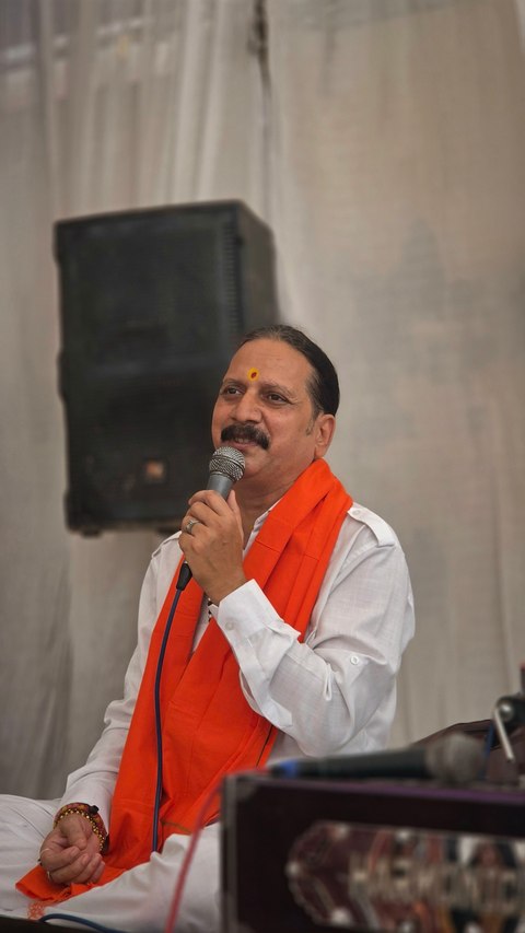 Prem Bhatnagar ji in white with a saffron stole, seated and singing into the microphone at a prayer meeting