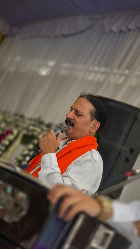 Prem Bhatnagar ji in white with a saffron stole, eyes closed as he sings a bhajan beside the flower-framed photo