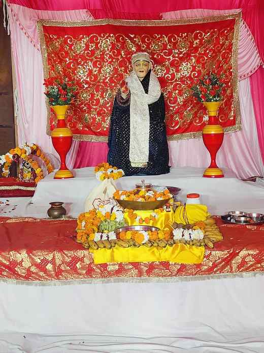 Sai Baba's murti in a white shawl before a red and gold backdrop, with marigolds, fruit and the jyot in front