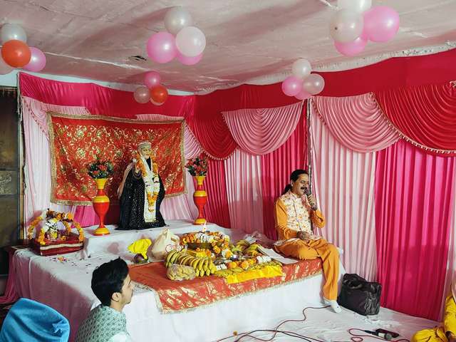 Prem Bhatnagar ji singing seated beside Sai Baba's darbar at a home Sai Sandhya, with fruit and flowers offered in front