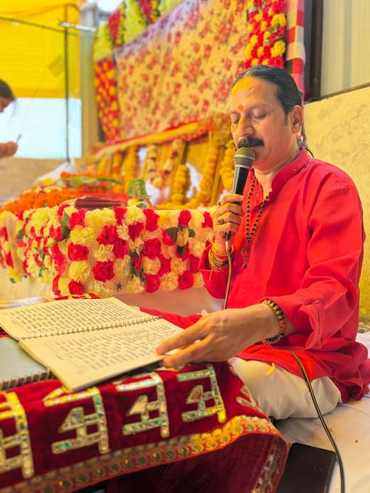 Prem Bhatnagar ji in a red kurta reading the Sunderkand into a microphone before the flower-decked darbar